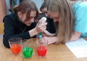 Na stole stoją trzy szklanki z wodą. W każdej szklance jest woda w innym kolorze. Kolejność szklanek od lewej strony: pomarańczowy, zielony, czerwony. W tle widać trzy uczennice, które nalewają ciecze w różnych kolorach do jednej szklanki.