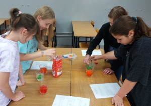 Uczennice przygotowują ciecze o różnych gęstościach. Wsypują cukier i barwniki do szklanek. W pierwszej szklance woda jest czerwona, w drugiej zielona a w trzeciej pomarańczowa.