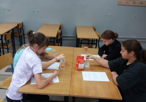 Przy stoliku siedzą cztery uczennice. Uważnie czytają instrukcję do doświadczenia. Na stole jest torebka z cukrem, dwie szklanki wypełnione wodą oraz kartki z instrukcją.