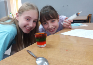 Uczennice są uśmiechnięte, ponieważ uzyskały ciecz o różnych warstwach. Przed dziewczynkami stoi szklanka wypełniona cieczą w trzech kolorach. Na dnie szklanki jest kolor pomarańczowy, wyżej zielony a nad nim czerwony.