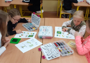 Cztery uczennice siedzą przy stolikach. Dziewczynki zapoznają się z instrukcją obrazkową. Łączą klocki według instrukcji.