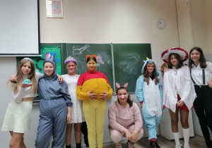 Grupa uczennic w kolorowych przebraniach stoi przy tablicy w szkolnej sali.