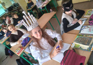 Uczniowie siedzą w ławkach w szkolnej sali. Dziewczynka w pierwszej ławce ma białą sukienkę i koronę na głowie, druga ma na twarzy maskę kota. W trzeciej ławce siedzi uczennica z rogami renifera na głowie. W dalszych ławkach siedzą pozostali uczniowie.