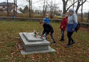 Trzech uczniów oraz nauczycielka podchodzą do obelisku na cmentarzu wojskowym z okresu I wojny światowej.