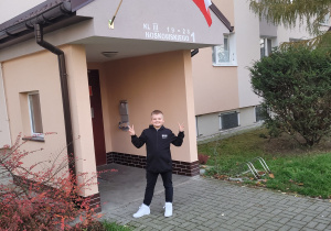 Uczeń stoi przed blokiem pokazując palcami znak Victorii, a nad nim znajduje się flaga biało-czerwona.