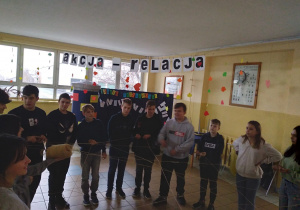 Grupa uczniów stoi na szkolnym korytarzu w kole, a w rękach trzymają rozwiniętą włóczkę wełny i podają ją między sobą, tworząc na środku sieć.