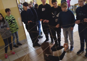 Kilkoro uczniów stoi na szkolnym korytarzu, w rękach trzymają grubą nitkę, podają ją między sobą i tworzą sieć. W środku siedzi dziewczynka i rozwija nitkę.