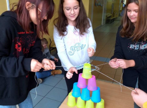 Uczennice budują wieżę z plastikowych kubeczków. Przenoszą kubki za pomocą sznurków przymocowanych do kubka.