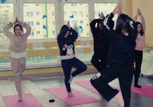 Uczennice stoją na matach w korytarzu szkolnym. Wykonują ćwiczenie psychofizyczne. Stoją na jednej nodze a drugą nogę zgiętą w kolanie opierają o udo, ręce unoszą nad głową.