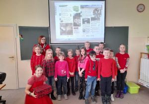 Grupa uczniów wraz z nauczycielką pozuje do zdjęcia w sali szkolnej, jenda dzeiwczynka sziedzi na podłodze, wszystcy mają czerwone ubrania.