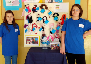 Wystawa laleczek UNICEF w korytarzu szkolnym. Na stoliku stoi tablica, do której są przypięte laleczki. Po obu stronach tablicy stoją uczennice w niebieskich koszulkach. Za wystawką wiszą plakaty Akcji Wszystkie Kolory Świata.