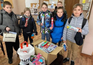 Sześcioro chłopców pozuje do zdjęcia w szkolnej sali lekcyjnej, w rękach każdy trzyma szczoteczkę lub pastę do zębów. Poniżej na stole stoją pudełka z zebranymi pastami i szczoteczkami do zębów.