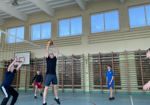 W sali gimnastycznej przez środek przewieszona jest siatka. Po obu strona siatki uczniowie odbijają piłkę sposobem górnym.