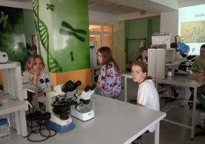 dwie dziewczyny i chłopiec w żółtej bluzie podpierający ręką głowę siedzą przy stole laboratoryjnym i przygotowują preparat do oglądania pod mikroskopem.