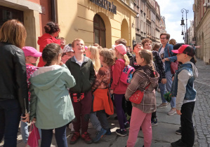 Grupa uczniów stoi na chodniku. Słuchają opowiadającej przewodniczki. Wokół nich znajdują się zabytkowe kamienice.