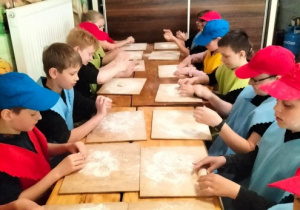 Uczniowie siedzą przy stole . Na głowie mają kolorowe czapeczki, ubrani są w fartuch. Na stole leżą deski kuchenne. Uczniowie lepią z ciasta bułki.