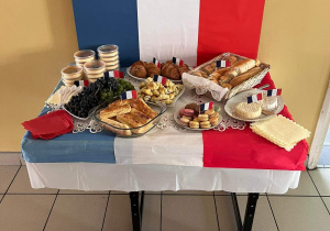 Widzimy stolik, na którym znajdują sie potrawy charakterystyczne dla Francji (winogrona, sery,makaroniki,bagietki,rogaliki).
