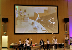 . Studenci robią doświadczenie z papierem. Na dużym ekranie wyświetlany jest pokaz doświadczenia z unoszącym się papierem.
