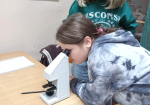 Zdjęcie przedstawia dwie uczennice pracujące z mikroskopem. Jedna z nich ogląda samodzielnie przygotowany preparat. Na stoliku leży kartka i długopis, na której uczennice będą zapisywały swoje obserwacje związane z preparatem.