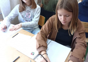 Zdjęcie przedstawia dwie uczennice siedzące w ławce. Dziewczyny mają przed sobą zeszyty, w których zapisują notatki z lekcji. Na ławce jest też kawałek cebuli, który służy do przygotowywania preparatów mikroskopowych.