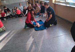 Uczniowie uczestniczą w ćwiczeniach z resuscytacji. Jedna z uczennic ćwiczy na fantomie, pod okiem strażaka.