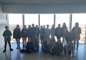 Uczniowie znajdują się na Sky Tower. Za nimi jest oszklona ściana,przez którą widać panoramę miasta.