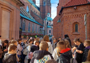 Uczniowie znajdują się na rynku we Wrocławiu. Stoją i słuchają pani przewodni. W tle widać piękną wrocławska katedrę.