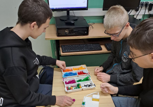 Uczniowie wybierają klocki Lego z pudełka. Budują konstrukcję, która jest na monitorze komputera.