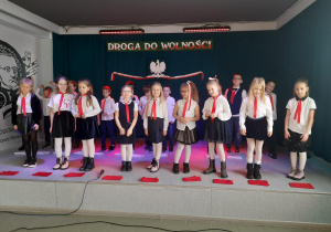 Grupa uczniów ubrana w biało – czarne stroje i z czerwonymi wstążkami na szyi stoi na scenie. W tle napis: Droga do wolności oraz ilustracja godła narodowego.