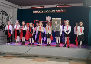 Na scenie stoi grupa dzieci odświętnie ubrana. Kilkoro z nich trzyma w dłoniach czerwone chusty. Jedna z dziewczynek trzyma w dłoniach portret Józefa Piłsudskiego.