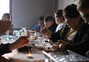 Uczniowie biorący udział w warsztatch chemicznych siedzą przy stole. Przy użyciu probówek i naczyń chemicznych pracują nad eksperymentami.