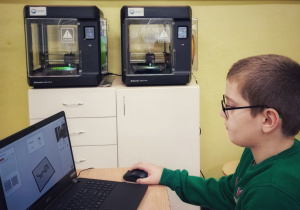 Uczeń siedzi przy stoliku obok drukarek 3D. Na stoliku jest laptop. Uczeń wyszukuje w Internecie model do drukowania i przesyła go do drukarki.