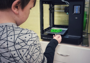 Sala lekcyjna, w której stoi drukarka 3D. Uczeń wyjmuje wydrukowany model z drukarki 3D.