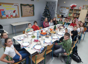 Uczniowie klasy siedzą wokół nakrytego stołu. Są na nim mandarynki, ciasto, cukierki, napoje.