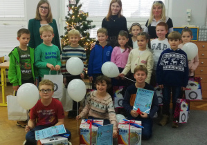 Uczniowie z wychowawczynią oraz pracownikami Wyższej Szkoly Kadr Menedżerskich w Koninie prezentują otrzymane nagrody i dyplomy. W ręku trzymają białe balony. W tle stoi udekorowana choinka.