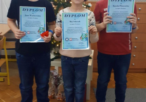 Troje uczniów, którzy zwyciężyli w konkursie prezentuje otrzymane dyplomy i zdobyte nagrody. W tle stoi udekorowana choinka.