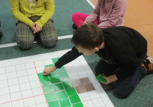 Uczeń siedzi na dywanie i układa wzór na macie do kodowania. Obok niego siedzą dwie uczennice.