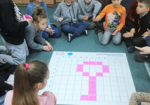 Grupa uczniów siedzi na dywanie wokół maty do kodowania. Na macie wzór klucza stworzony przez uczniów.