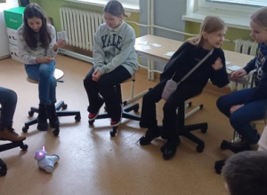 Dzieci siedzą w kręgu na krzesełkach. Na podłodze porusza się robot.