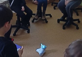 Uczniowie siedzą w kręgu na krzesełkach. Jeden z chłopców steruje robotem na telefonie. Robot porusza się po sali.