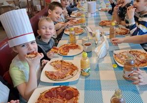 Wzdłuż długiego stołu, po obu stronach siedzą dzieci. Przed dziećmi stoją na stole talerze z upieczoną pizzą. Dzieci jedzą.
