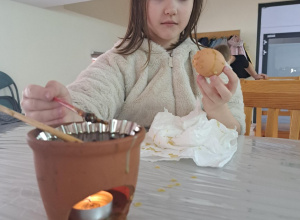 Dziewczynka siedzi za stołem i maluje jajko. Na pierwszym planie na stole stoi pojemnik z woskiem. Pojemnik ma otwór, przez który widać palący się podgrzewacz.