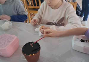 Dziewczynka siedzi za stołem i maluje jajko. Na pierwszym planie na stole stoi pojemnik z woskiem. Na drugim planie siedzą dzieci wokół stołu.