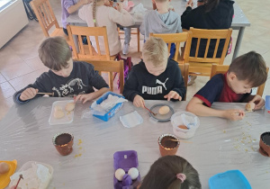 Widok z góry na stół. Przy stole siedzą dzieci. Dzieci malują jajka. Na stole stoją pojemniki z woskiem. Na drugim planie kolejny stół, wokół którego siedzi kolejna grupa dzieci.