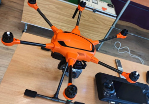 Na stole stoi duży dron w kolorze pomarańczowym. Obok niego leży urządzenie sterujące.