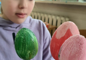 Uczennica trzyma w dłoniach trzy styropianowe jajka pomalowane kolorowymi farbami, umieszczone na drewnianych patyczkach.