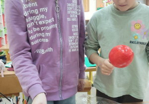 Dwie uczennice trzymają w dłoniach pomalowane jajka umieszczone na drewnianych patykach. Jedna z nich zanurza jajko w pojemniku z cukrem.