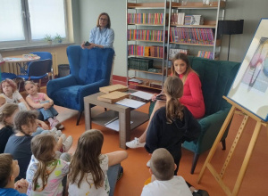 W pomieszczeniu bibliotecznym tyłem siedzi grupa dzieci. W fotelu siedzi pracownik biblioteki, obok stoi nauczyciel. W tle regały z książkami.