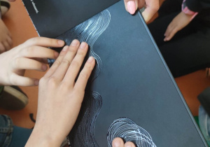 Uczniowie dotykają palcami książki przeznaczonej dla osób niedowidzących. Książka jest czarna i ma specjalne fakturowania.
