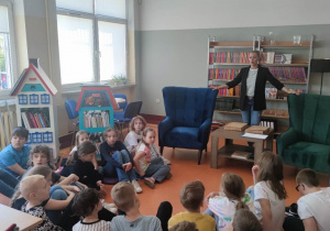 W pomieszczeniu bibliotecznym tyłem siedzi grupa dzieci. W fotelu siedzi pracownik biblioteki, W tle regały z książkami.